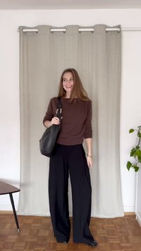 Pulover tricotat maro cu tiv elastic, asortat cu pantaloni negri evazați. Geantă din piele neagră cu curea, așezată pe un fundal de perdea deschisă.