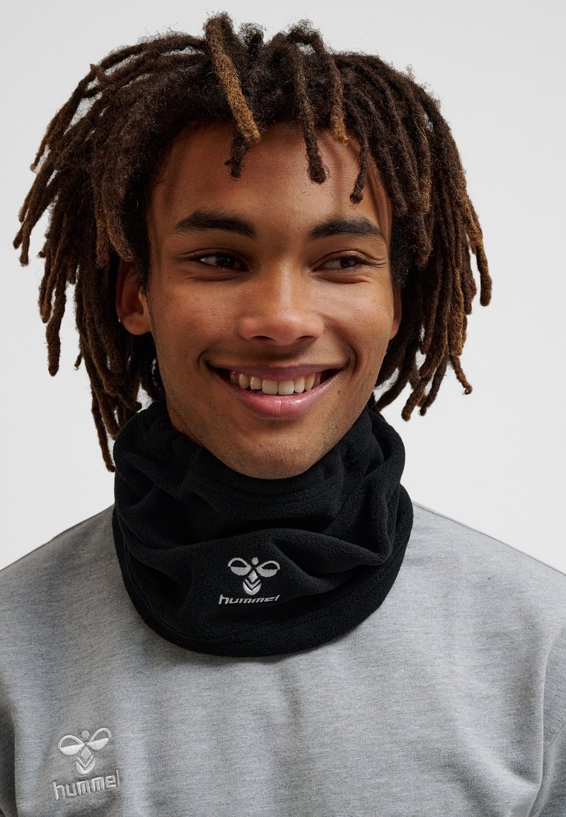 Smilende person med mellemlange dreadlocks iført en sort fleece halsvarmer og en lysegrå skjorte, begge med hummel-logoer.