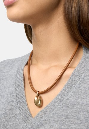 Brązowy naszyjnik z rzemienia skórzanego z złotym metalicznym wisiorkiem o organicznym kształcie i gładkiej fakturze, leżący na szarym swetrze.