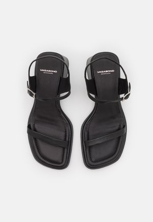 Nainen, joka pukeutunut mustaan hihattomaan toppiin, mustiin nahkahousuihin ja mustiin korkokenkäsandaaleihin, istuu metallikehyksisellä punotulla rottinkituolilla.