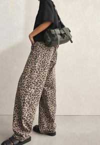 Beżowo-czarne szerokie spodnie w panterkę, zestawione z luźną czarną koszulą i zieloną torebką na ramię. Czarne sandały.