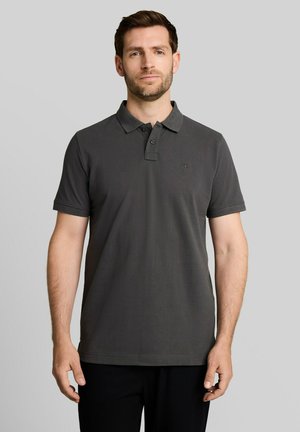Man met kort bruin haar en baard, gekleed in een donkergrijs poloshirt met korte mouwen, staand voor een effen lichtgrijze achtergrond.