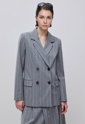 Giovane donna che indossa un blazer grigio a doppiopetto a righe sottili e pantaloni abbinati, occhiali da sole appoggiati sulla testa, sfondo neutro.