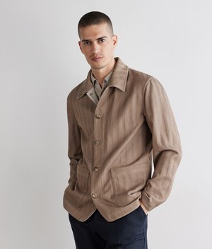 Mand med kort hår iført en beige knappet jakke over en skjorte og mørke bukser, stående mod en almindelig hvid væg.