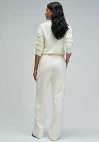 Femme aux longs cheveux noirs portant un haut à manches longues blanc cassé et un pantalon assorti, se tenant les mains dans les poches, de dos contre un fond gris.