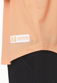 T-shirt a rete pesca con etichetta bianca recante la scritta "PEGADOR NEVER TOO LATE" in testo dorato, abbinata a pantaloni neri con tasche laterali.
