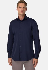 Muž v priliehavých tmavomodrých dlhých rukávoch košeľa s golierom a svetlosivých nohaviciach, stojí proti jednoduchej pozadí.