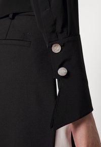 Rukáv černého na míru šitého saka má stříbrné knoflíky, textura látky je hladká s jemným leskem a nad kapsou je vyšívané logo.