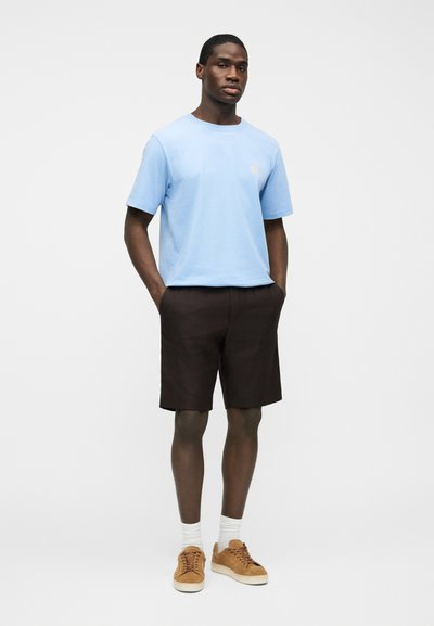 Άνδρας που στέκεται με τα χέρια στις τσέπες, φοράει ανοιχτό μπλε μπλουζάκι, σκούρα σορτς, λευκές κάλτσες και ταμπά αθλητικά παπούτσια, σε απλό λευκό φόντο.