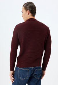 Bordeaux gebreide trui met een textuurpatroon, lange mouwen en geribbelde manchetten. Gecombineerd met blauwe denim jeans. Zicht van achteren.