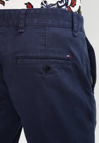 Marlové modré bavlněné kalhoty se zadní kapsou, která má černý knoflík a malou červenou, bílou a modrou logovou ozdobu poblíž uzávěru kapsy.