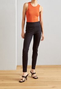 Orange ribbad tanktop kombinerad med högmidjade svarta byxor och svarta klacksandaler. Trägolv och neutral vägg som bakgrund.