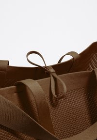 Borsa tote in rete marrone con ampie spalline in tessuto. I dettagli notevoli includono un design annodato sulle spalline e un corpo liscio e testurizzato.
