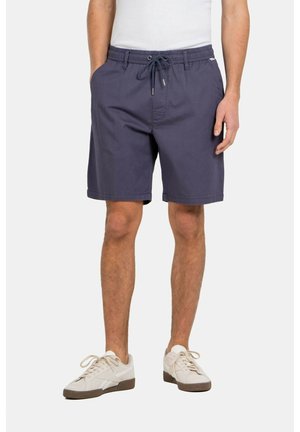 Mann trägt marineblaue Shorts mit Kordelzug, weiße Sneakers und ein weißes Hemd, steht vor einem schlichten hellen Hintergrund.