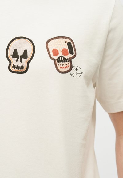 Δύο κεντημένες μπαλώματα με κρανία σε μαύρο, πορτοκαλί και μπεζ σε ένα λευκό πουκάμισο, με το λογότυπο "PS Paul Smith" κοντά στο δεξί κρανίο.