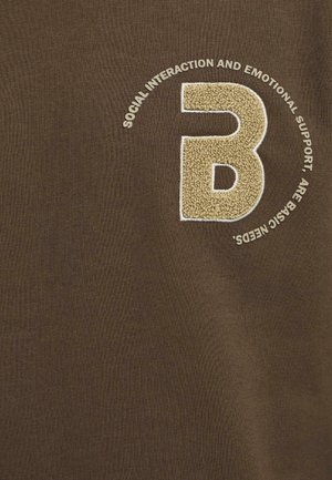 Smeđi pamučni majica s vezenom bež letters "B" i okolnim tekstom u kružnom uzorku. Glatka tekstura, okrugli izrez.