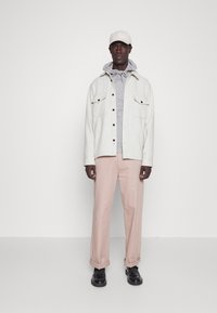 Veste en mélange de laine gris clair avec deux poches poitrine, superposée à un hoodie gris. Un pantalon rose pâle et une casquette beige complètent le look. Chaussures noires.