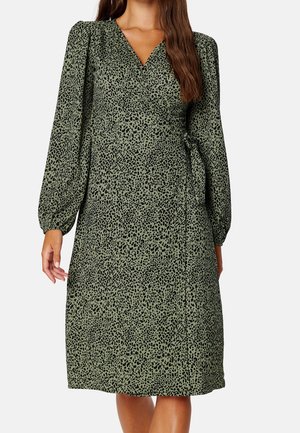 Femme portant une robe portefeuille vert olive longueur genou avec un motif noir tacheté et de longues manches bouffantes.