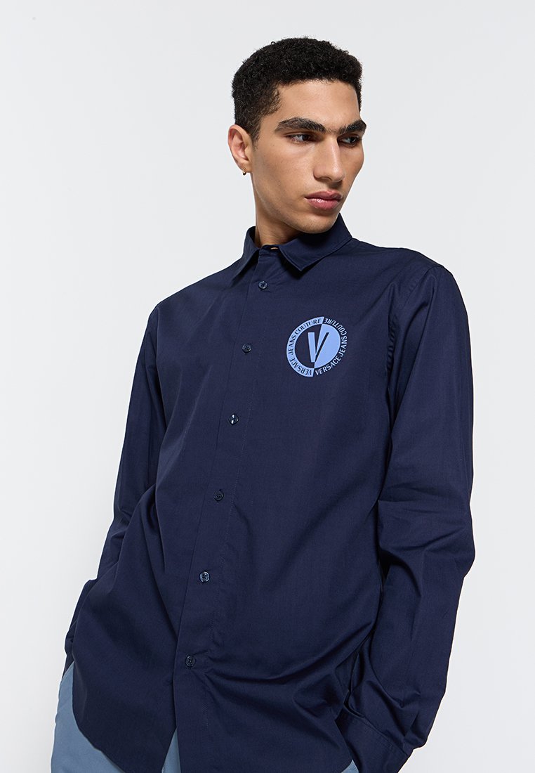 Chemise boutonnée bleu marine en tissu lisse, avec un logo rond bleu clair sur la poitrine gauche, un col classique et des manches longues.