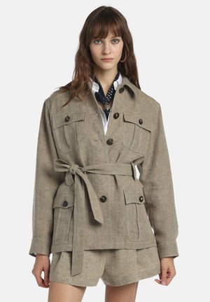 Kvinde iført beige jakke med bælte og fire knapstolperede lommer samt matchende shorts, lagdelt over hvid skjorte og marineblå tørklæde med polkaprikker.