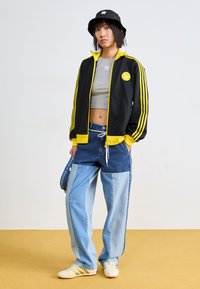 Černá sportovní bunda s žlutými akcenty a logem usmívající se tváře, šedý crop top a džíny s patchworkovým designem. Bílé boty se žlutými pruhy a černý baret.