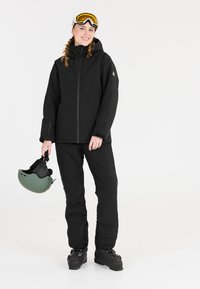 Schwarze Skijacke mit Kapuze, Reißverschluss und entspannter Passform, kombiniert mit passenden Hosen. Tr�ägt einen grünen Skihelm und schwarze Skistiefel.