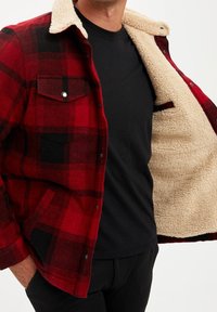 Man in een rood en zwart geruit jasje met beige fleece voering, toont de open binnenkant van het jasje over een zwart shirt.