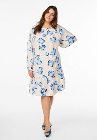 Robe crème avec motif floral bleu, manches longues, encolure ronde et ourlet froncé. Modèle portant des chaussures à talons hauts bleues.