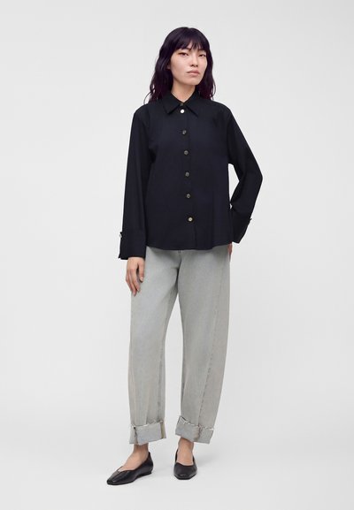 Femme debout portant une chemise noire à manches longues boutonnée, un pantalon gris clair retroussé et des chaussures plates noires sur un fond uni.