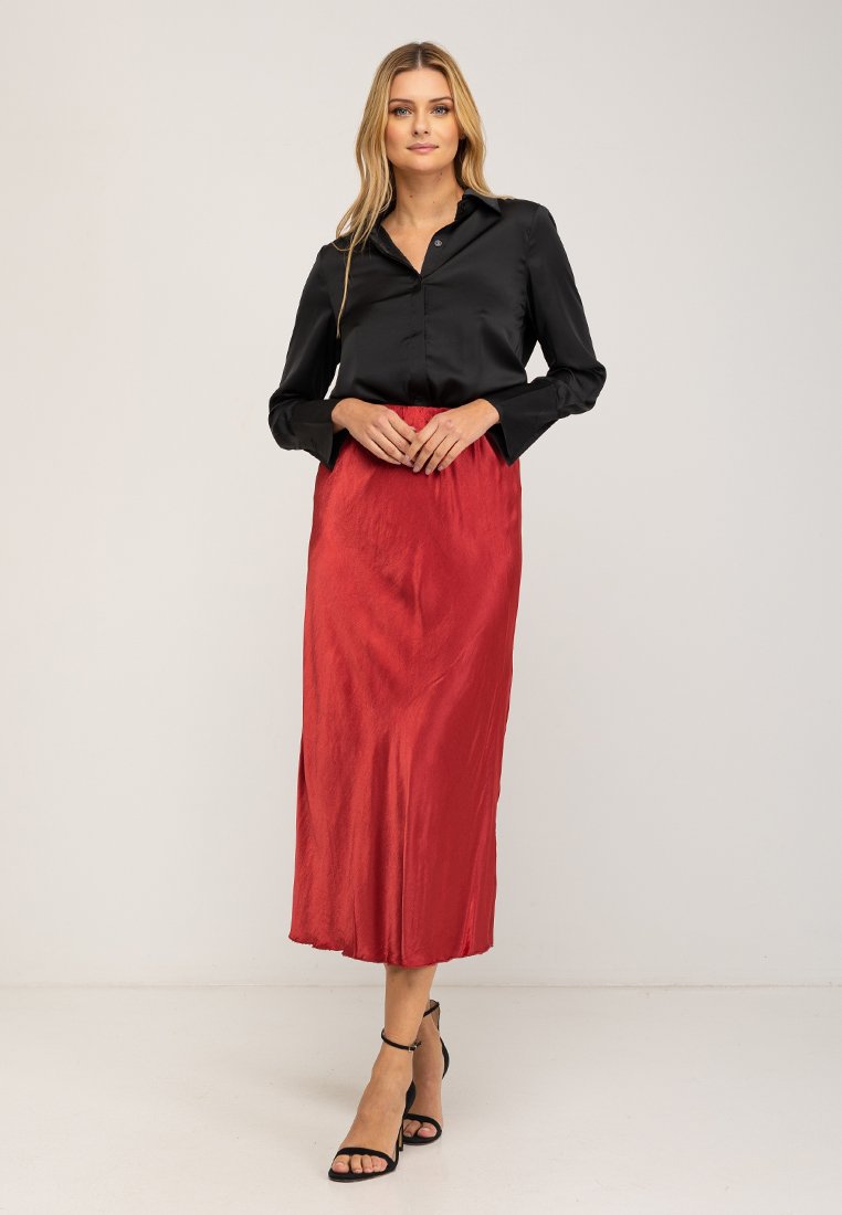 Zwarte satijnen blouse met lange mouwen, gecombineerd met een knielange, vloeiende rode rok met een subtiele glans, aangevuld met strappy zwarte sandalen.