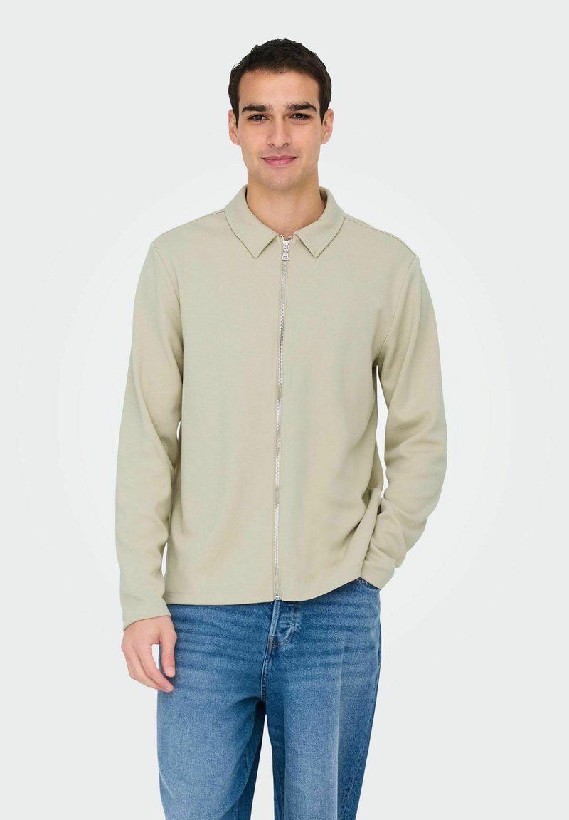 Light beige zip-up jacket with a collar, long sleeves, and a smooth texture, paired with blue jeans. Simple design without patterns.