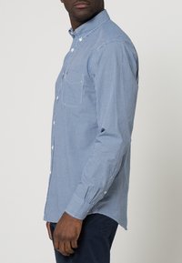 Camisa de botón en un patrón de cuadros azul y blanco, con un collar clásico, mangas largas y un dobladillo curvado. Tejido suave y de punto.