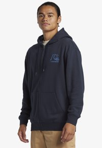 Námornícky zipsový mikina vyrobená z jemného materiálu má prednú klokankovú vrecko, nastaviteľnú kapucňu so šnúrkou a modrú potlačenú značku na hrudi.