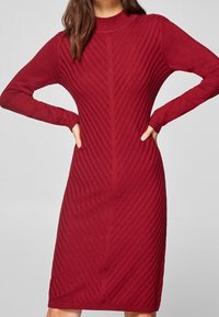 Rotes Strickkleid mit langen Ärmeln, vertikaler Ripptextur, Rundhalsausschnitt und tailliertem Schnitt. Midi-Länge, minimalistische Details.
