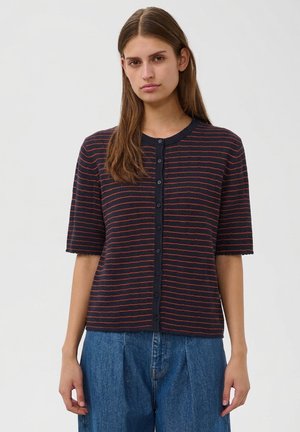 Jeune femme aux longs cheveux bruns porte un pull bleu marine à manches courtes boutonné avec de fines rayures horizontales rouges et un jean bleu.