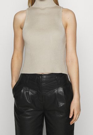 Personne portant un haut sans manches beige à col roulé et un pantalon noir en cuir taille haute, debout les mains dans les poches devant un fond uni.