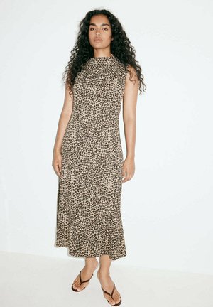 Femme aux cheveux bouclés portant une robe midi sans manches à motif léopard et des tongs noires, debout devant un fond clair uni.