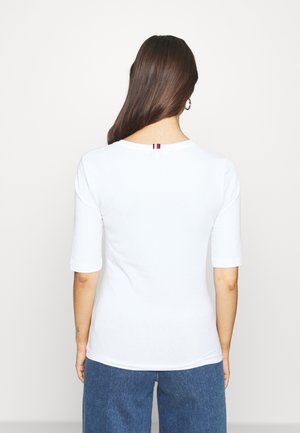 Vrouw met lang bruin haar die een wit T-shirt met korte mouwen draagt met een klein rood, wit en blauw label aan de achterkant van de halslijn en blauwe spijkerbroek.