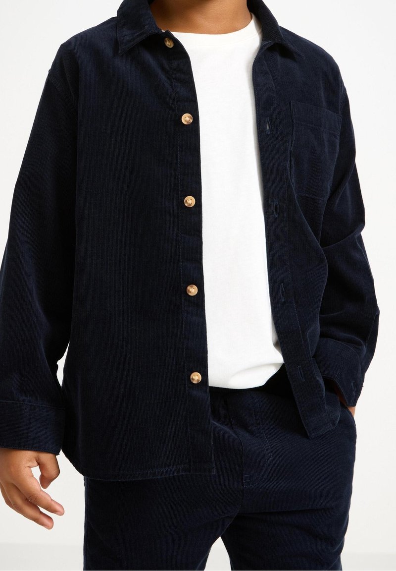 Enfant portant une chemise en velours côtelé bleu marine boutonnée sur un t-shirt blanc, avec un pantalon en velours côtelé bleu marine assorti.