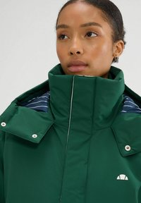 Zielona, ocieplana kurtka z wysokim kołnierzem, srebrnymi zatrzaskami i paskowanym wnętrzem. Małe logo z przodu. Gładka tekstura.