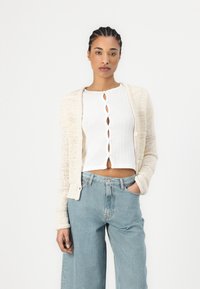 Light cream knitted cardigan over a white ribbed top with a wavy button placket, paired with loose light blue jeans.