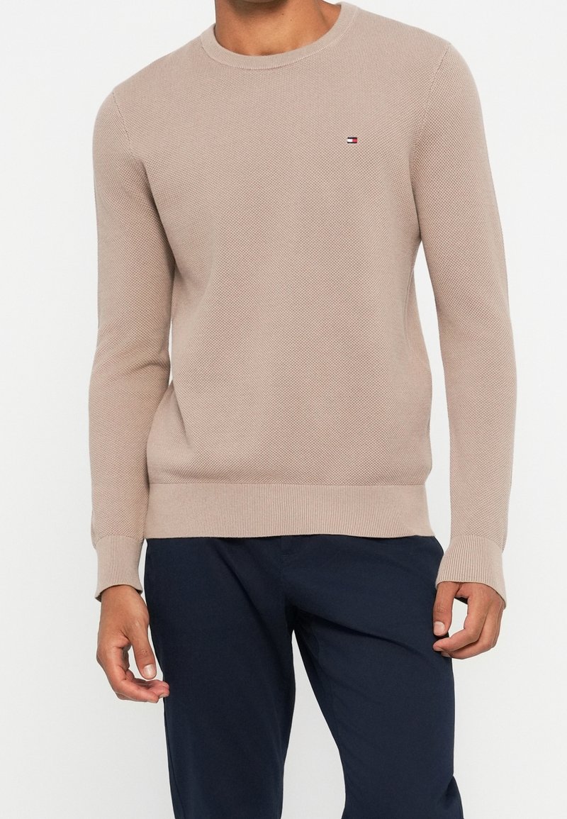 Homme portant un pull beige à manches longues avec un petit logo sur la poitrine et un pantalon bleu foncé, debout devant un fond blanc uni.