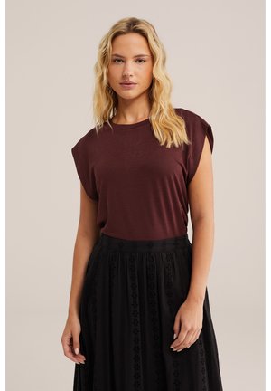 Šviesiaplaukė moteris su bangeliais plaukais, dėvinti rankovių neturintį bordo viršų ir juodą aukštos liemens sijoną su nėrinių raštais, stovinti priešais paprastą foną.