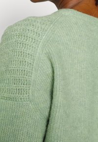 Gros plan d'une personne portant un pull vert clair doux et texturé avec des motifs de tricot variés sur l'épaule et la manche.