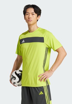 Un maillot de sport vert vif avec des accents noirs et trois bandes verticales. Le joueur tient un ballon de football noir et blanc.