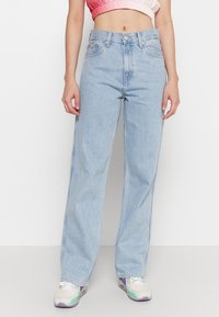 Jeans de pierna ancha en azul claro con cintura alta, confeccionados en denim. Presentan un cierre de botón, cinco bolsillos y un sutil desgastado. Combinados con zapatillas.