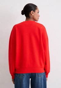 Czerwony, oversize sweter z okrągłym dekoltem, opadającymi ramionami i ściągaczami, w zestawieniu z niebieskimi dżinsami. Miękka faktura.