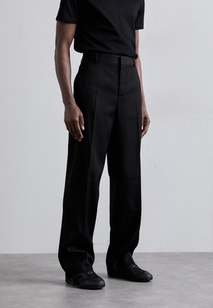 Black wide-leg trousers with a smooth texture, pleats at the front, paired with a fitted black t-shirt and black sneakers.