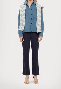 Camicetta azzurro chiaro con taschino sul petto, sovrapposta a un gilet senza maniche a righe. Abbinata a pantaloni svasati blu scuro e sandali.