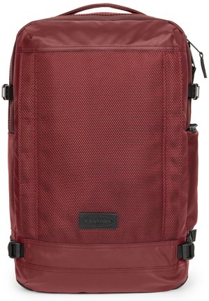 Red fabric backpack with a textured surface, flat top, side pocket, adjustable straps, and a brand logo patch on the front.